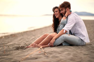 Genç ve güzel bir çift kumlu sahilde oturuyor, eğleniyor, gülümsüyor ve sarılıyorlar. Aşk, rahatlık, rahatlama, şefkat konsepti