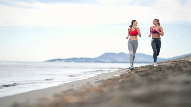 İki kadın sahilde birlikte koşuyor ve antrenman yapıyor.