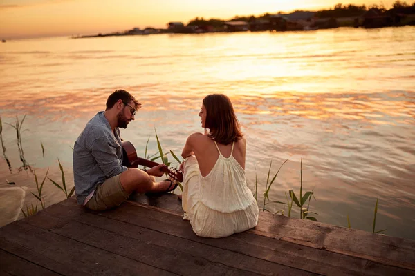 小伙子在湖面上为他的女朋友弹奏吉他 夕阳西下 — 图库照片