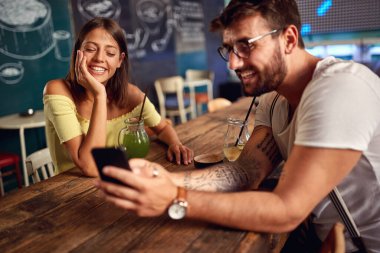 Genç çift bir kafede içki ve cep telefonuyla eğleniyor.