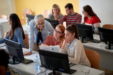 Öğrenciler, üniversitenin bilgisayar sınıfındaki bir bilgilendirme dersinde kadın profesörden yardım istiyorlar.