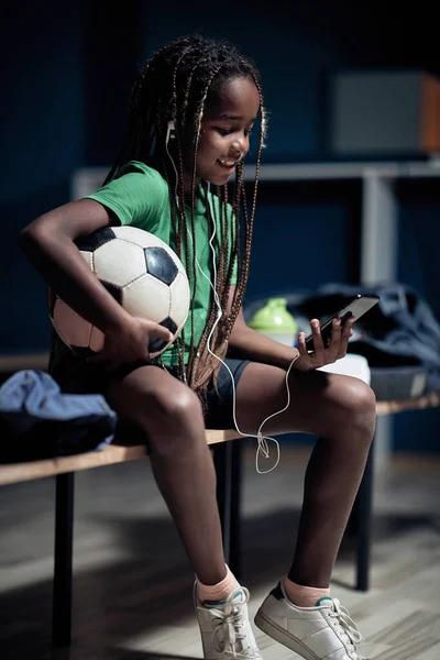一个小足球运动员正在更衣室里等待训练 他喜欢手机上的内容 — 图库照片