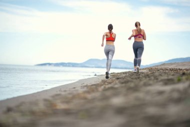 Genç kızlar güzel bir havada deniz kenarında koşuyor.
