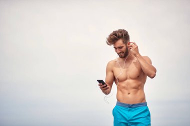 Kumsalda genç yakışıklı bir adam güneşli bir havada akıllı telefonuyla müzik dinliyor.