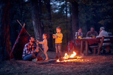 Babalar ve çocuklar güzel bir sonbahar günbatımında ormanda kamp ateşi etrafında oynuyorlar.