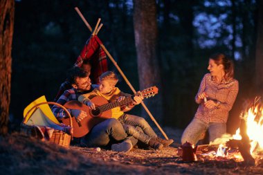 Genç ve mutlu bir aile ormanda güzel bir gecede gitarın ve kamp ateşinin tadını çıkarıyor.