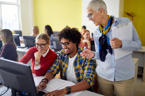 A female professor following work of students at the informatics lecture in the university computer classroom. Smart young people study at the college. Education, college, university, learning and multiethnic people concep