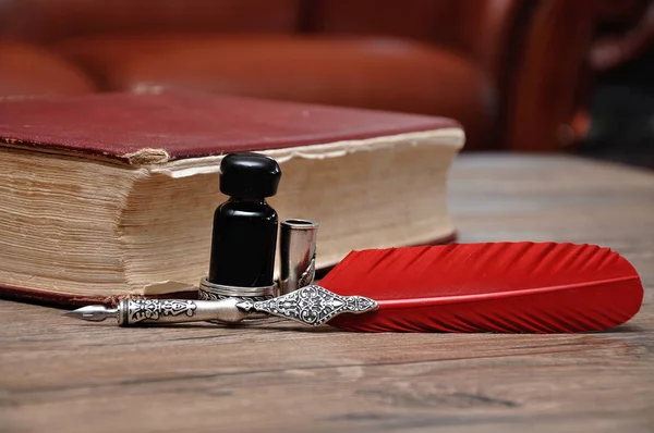 Ahşap bir masanın üzerinde kırmızı bir kitapla bir şişe mürekkep olan eski bir kalem.