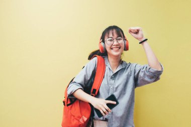 Teen lifestyle concept. Happy pretty hipster girl in earphones listening to music and dancing and holding mobile phone on bright yellow background. clipart