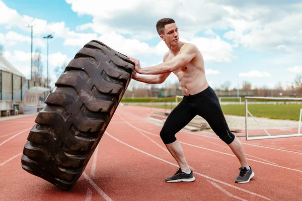Young muscular man lifting up huge tire for training muscles on stadium