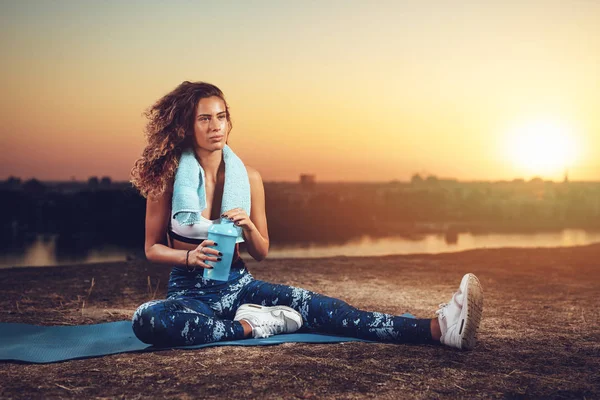Young fitness woman resting after hard training by river at sunset