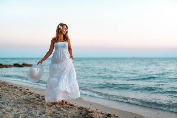 穿着白色礼服的年轻微笑的妇女与夏天帽子摆在海滩上 — 图库照片
