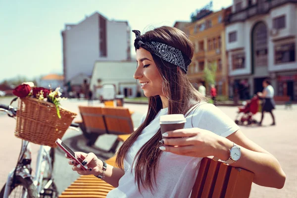 young woman with bike with flower basket using smartphone and drinking coffee