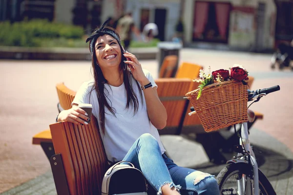 young woman with bike with flower basket using smartphone and drinking coffee