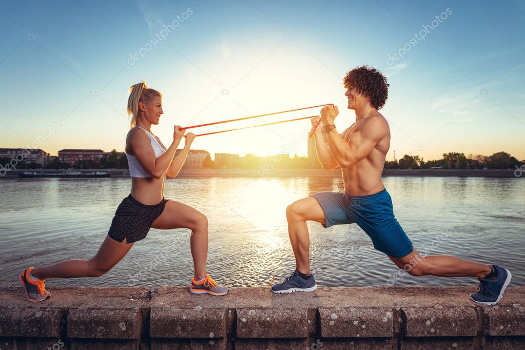 Young fitness couple doing workout with rubber band by river at sunset 