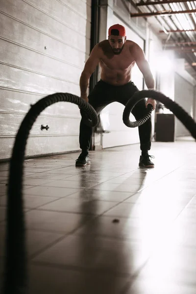 muscular young man exercising with ropes in garage gym