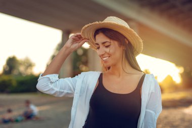 Hasır şapkalı günbatımı zaman nehir kıyısında yürüyen genç gülümseyen kadın