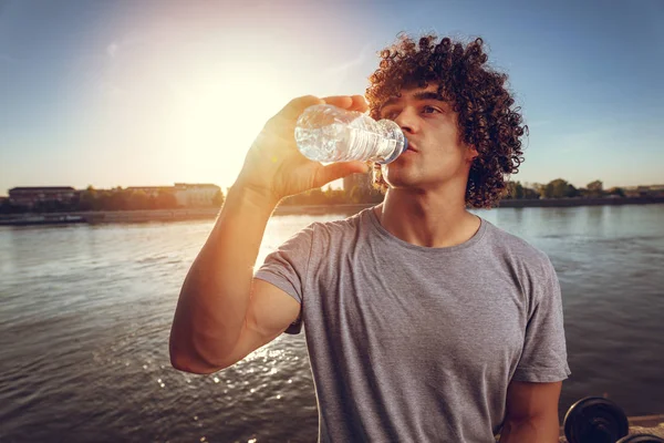 Young sport guy resting after hard training by river at sunset and drinking water