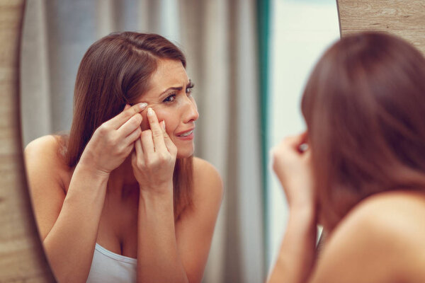 Young woman looking on pimple on cheek in mirror and frowning