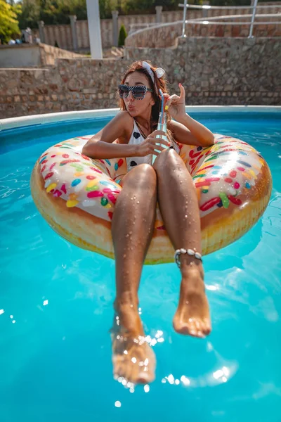 young woman with cocktail swimming on inflatable ring donut in swimming pool