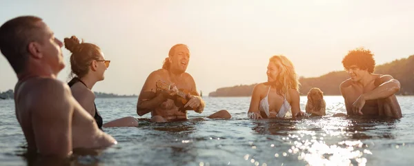 Grupo Amigos Felices Sentados Agua Hombre Jugando Ukelele Atardecer —  Fotos de Stock