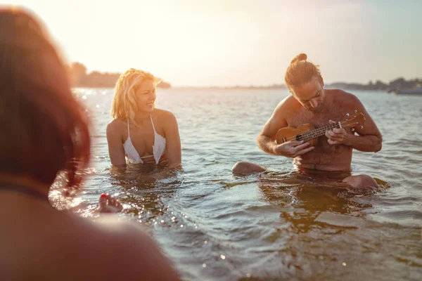 朋友坐在水和人演奏四弦琴在 Sunsest — 图库照片