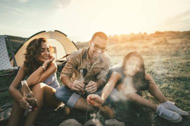 Mutlu genç arkadaşlar Dağı'nda bir gün batımının keyfini çıkarın. Onlar gülüyor ve çadır yakınındaki şenlik ateşi tarafından dışarı asılı.