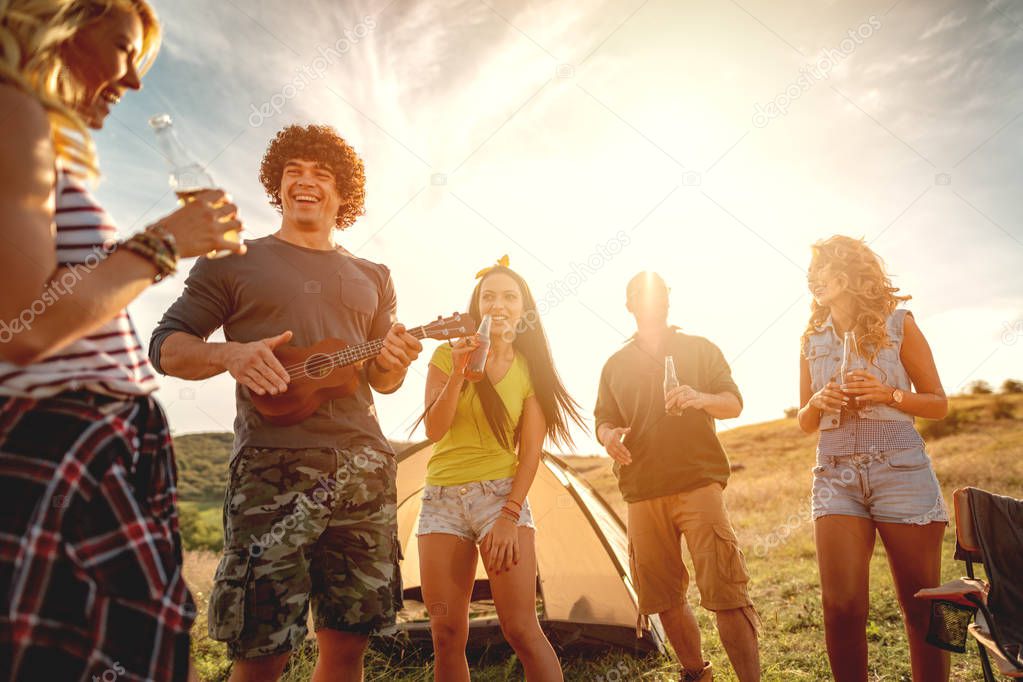 Young people have a good time in camp in nature. They're resting, laughing and singing with music from ukulele, happy to be together.