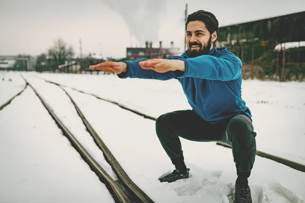 Active young man crouching and doing exercises in public place on winter training