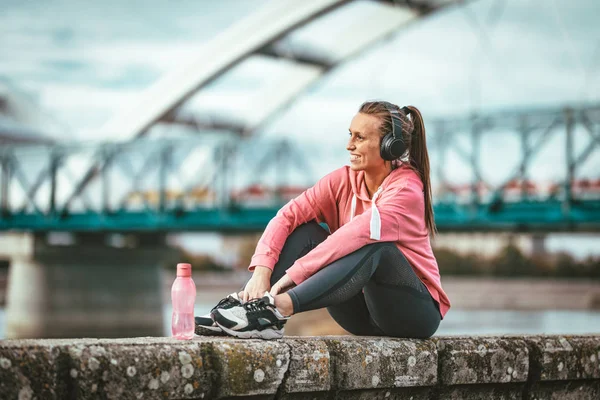 Young fitness woman resting after hard training on wall by river bridge