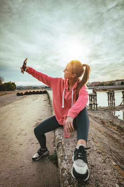 Young fitness woman resting after hard training on wall by river bridge and taking selfie with smartphone