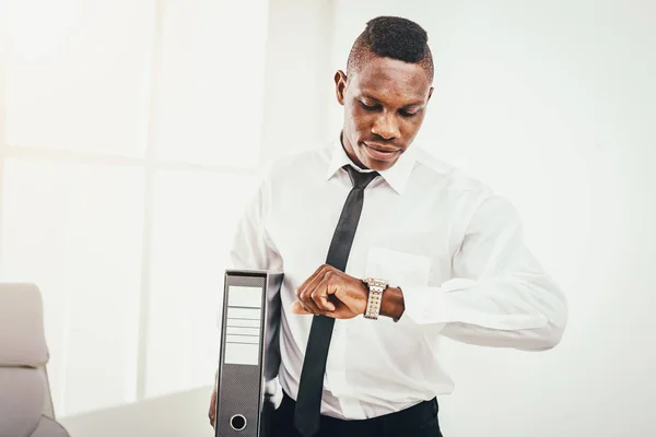 Successful African businessman looking at watch and holding folder in modern office