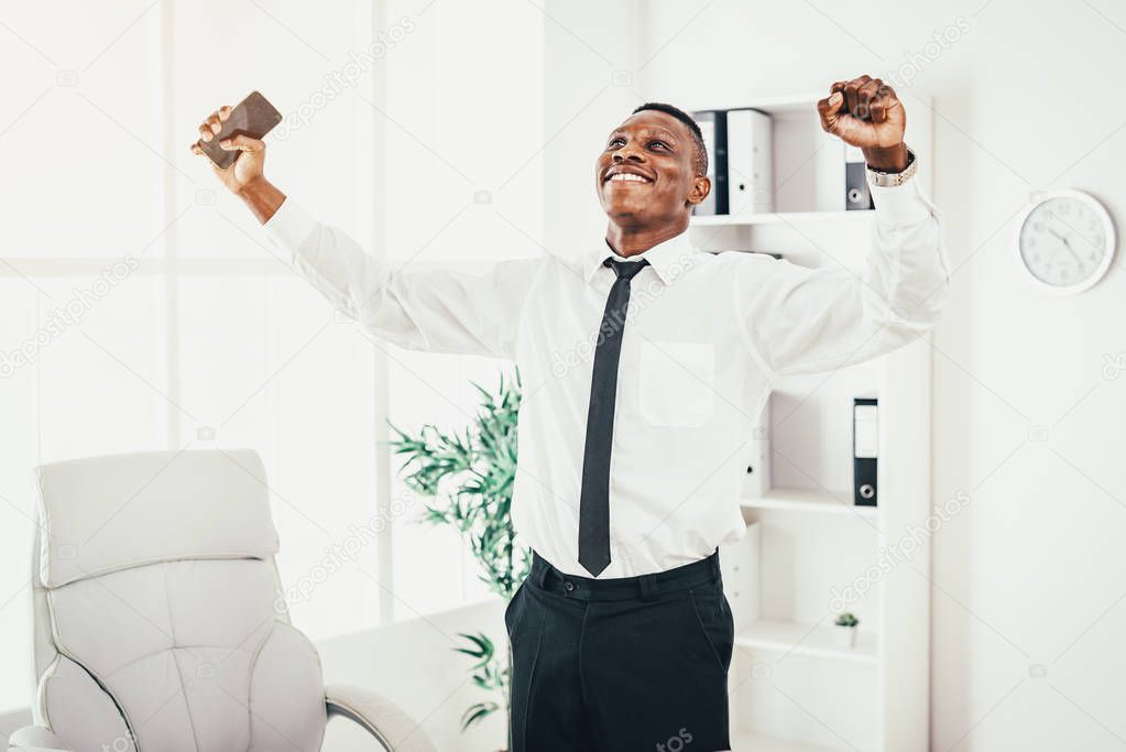 Successful African businessman celebrating success with raised arms in modern office