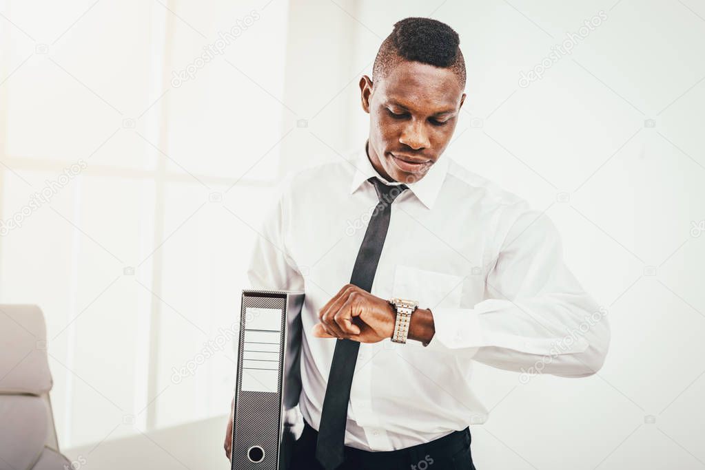 Successful African businessman looking at watch and holding folder in modern office