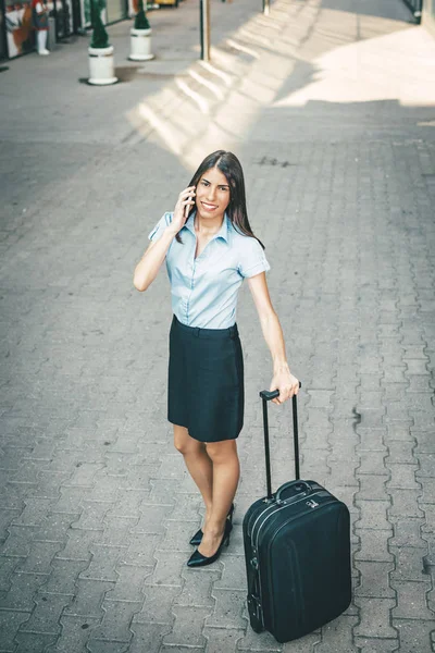 Young businesswoman standing  with suitcase and talking on smartphone