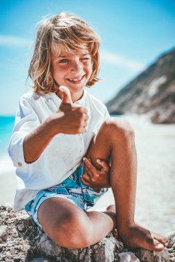 plaj ve kameraya bakarak deniz kaya üzerinde oturan çocuk gülümseyen 