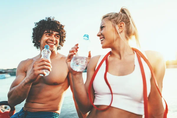 Young fitness couple resting after hard training by river at sunset drinking water