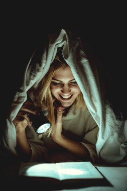 Genç kadın kapsayan ve el feneri tutan yatağının altında kitap okuma