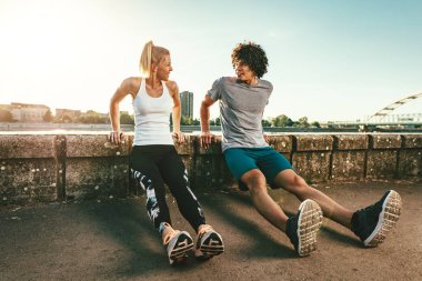Young happy smiling couple runners are training outdoors doing stretching exercise on the wall by the river.