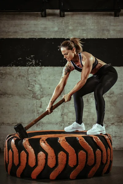 Young muscular woman hitting wheel tire with hammer on training at gym