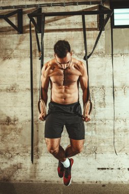 Genç kaslı adam spor salonunda jimnastik halkaları üzerinde egzersiz yapıyor.