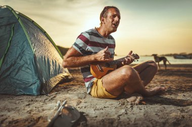 nehir kenarında oturan ve gün batımında ukulele oynarken müzisyen