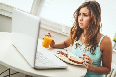 Genç bir kadın kahvaltı yapıyor ve dizüstü bilgisayarla çalışıyor.