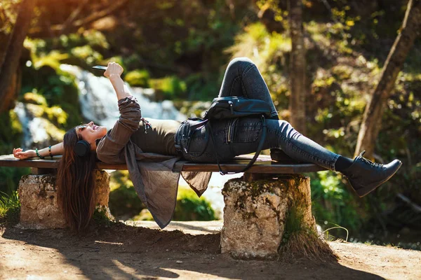 smiling young woman lying down on bench and listening music in mountains