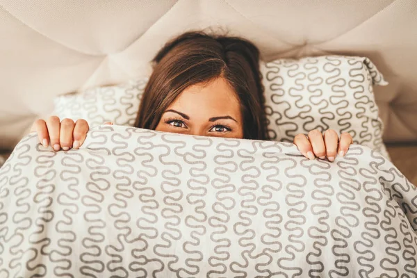 Young woman hiding over blanket in bed