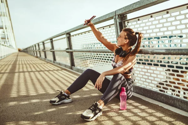 Young fitness woman resting after hard training on river bridge and taking selfie with smartphone
