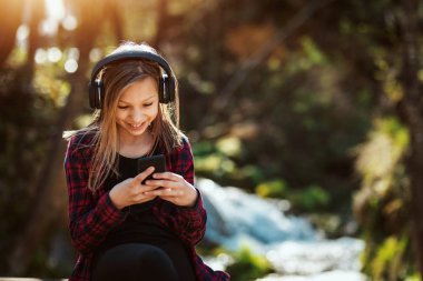 Gülümseyen küçük kız akıllı telefondan müzik dinliyor ve dağlarda manzaranın tadını çıkarıyor