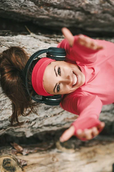 Young happy female runner lying on wooden log and listening to music after running in forest