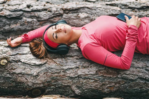 Young happy female runner lying on wooden log and listening to music after running in forest
