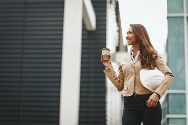 Genç kadın mimar kahve içme ve kentsel şehir arka planda elinde kask tutarak
